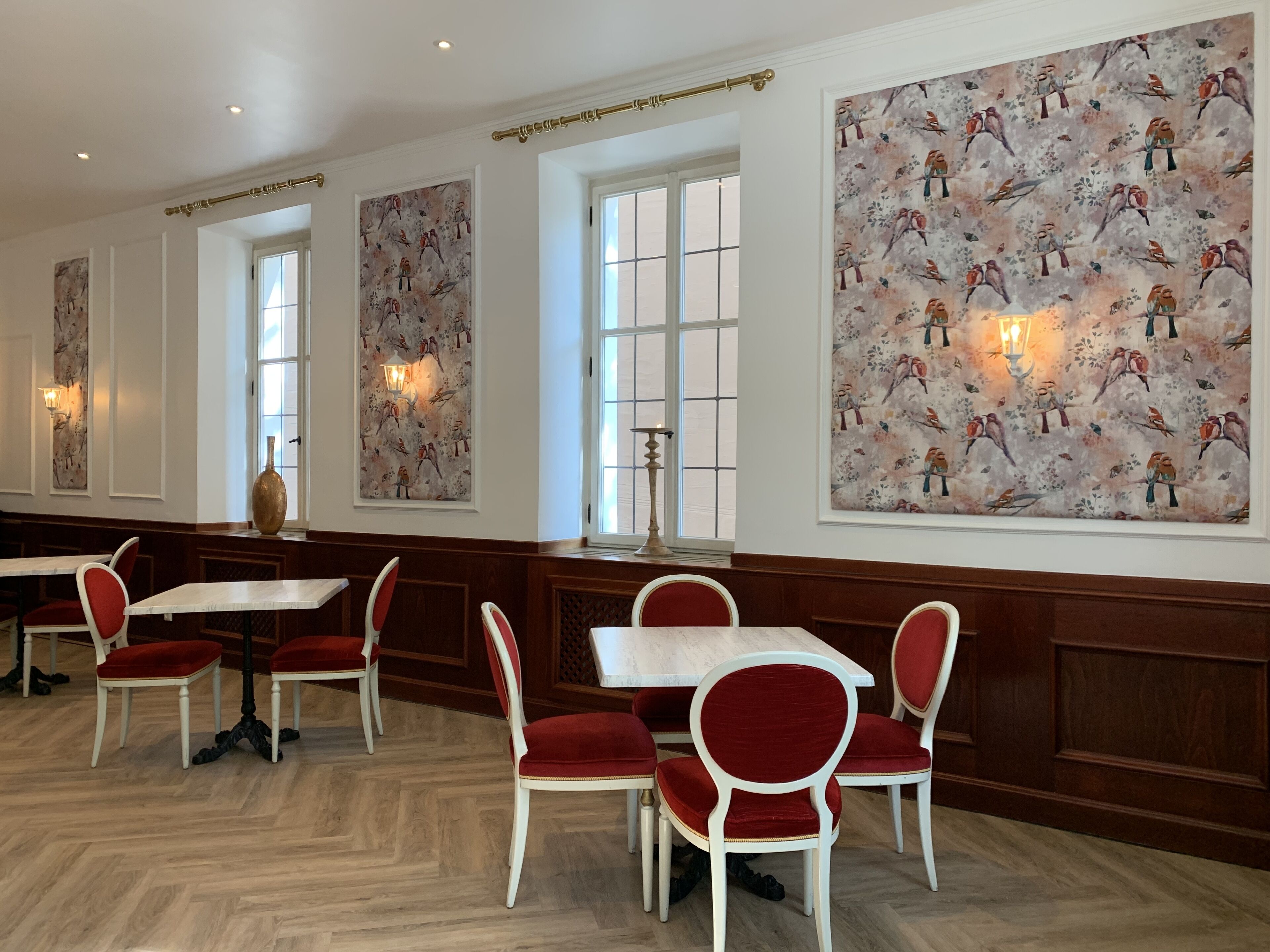 A dining area with white tables and red upholstered chairs, large windows, and wall art depicting birds.
