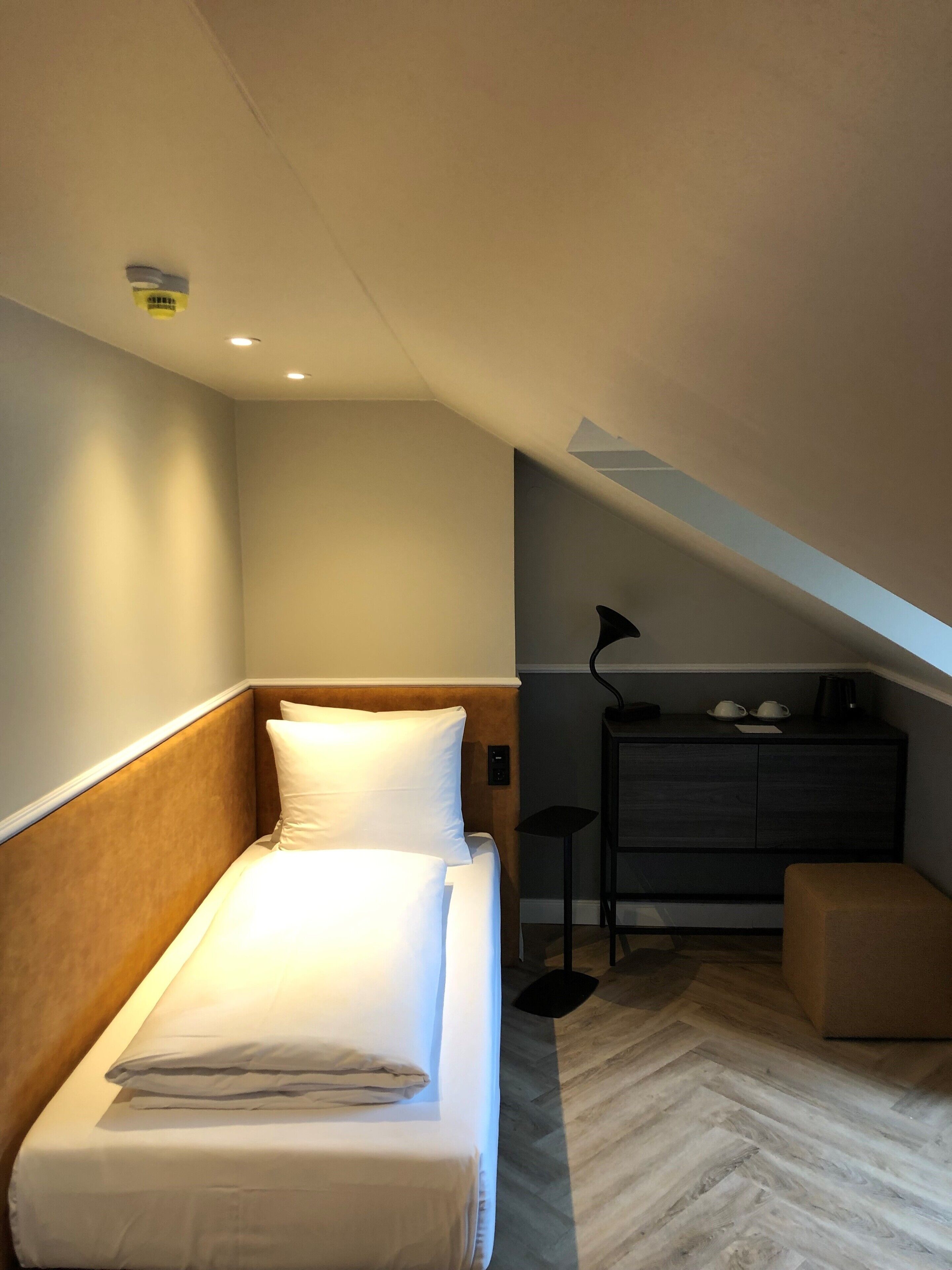 A neatly made bed in a small, modern room with a herringbone-patterned floor, a desk with a chair, and a small ottoman.