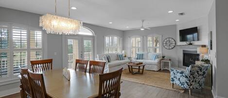 A spacious living room with a dining area, large windows, a fireplace, and a ceiling fan.