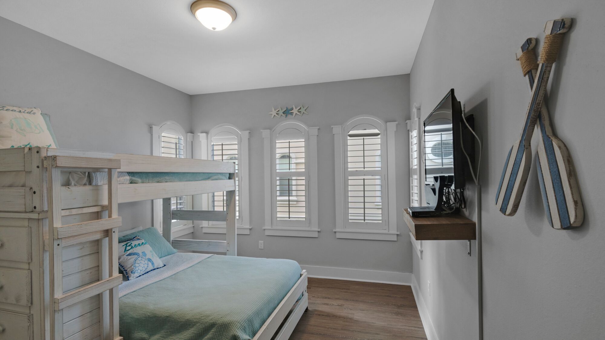 A bedroom with a bunk bed, a desk, and a mirror. There are three windows with shutters.
