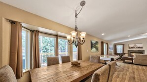 A spacious living room with a wooden dining table, comfortable seating, a fireplace, and large windows offering a view of the outdoors.