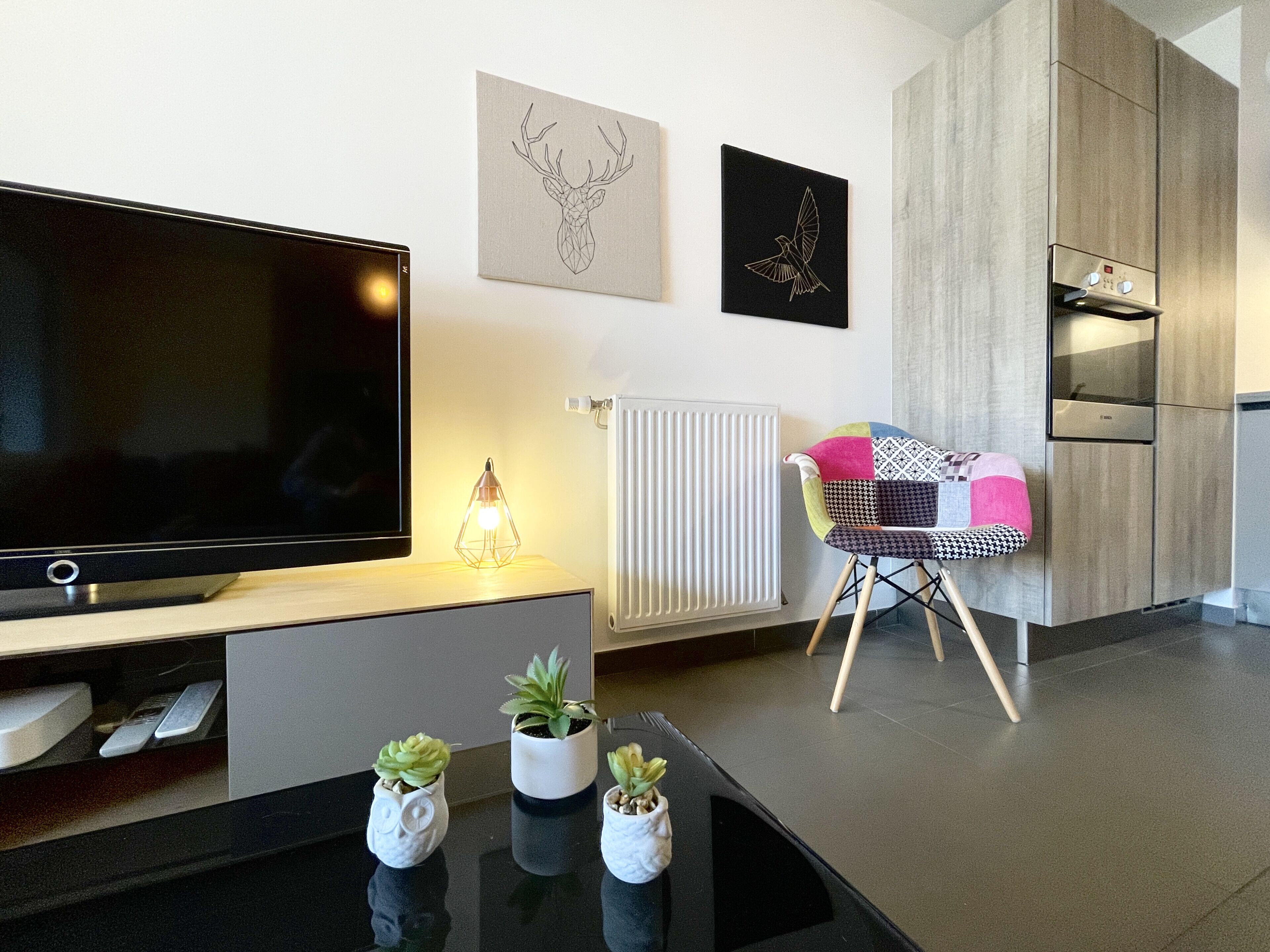 A modern living room with a flat-screen TV, a radiator, a wooden cabinet, and a chair with a colorful cushion.