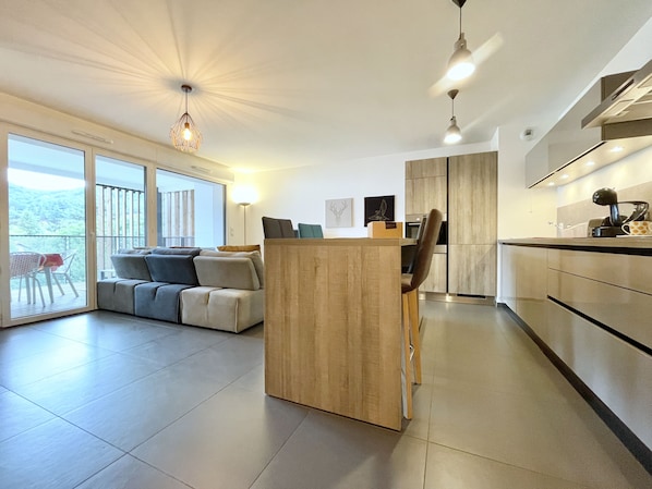 Eine moderne Küche mit einer Holztischinsel, einem Essbereich mit einer Couch und Blick durch große Schiebetüren auf einen Balkon.