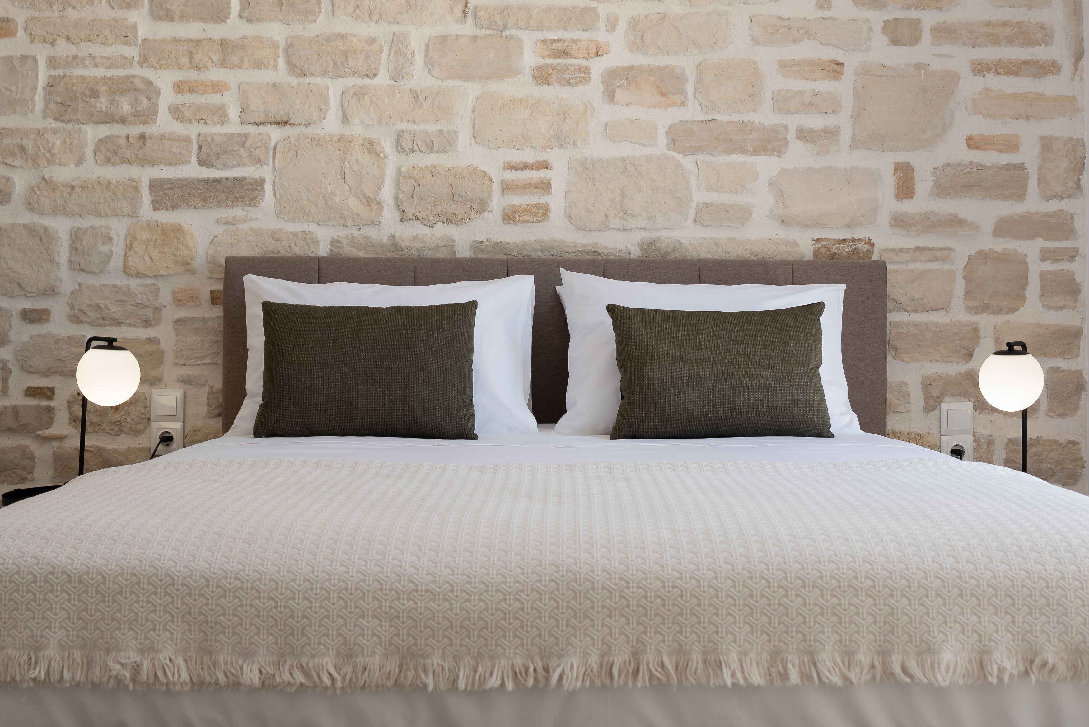 A bed with a white bedspread and two pillows, one white and one green, against a stone wall.