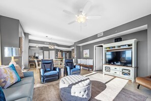 Un salon moderne avec un canapé gris, des fauteuils bleus, un pouf gris, un meuble de rangement blanc avec un téléviseur et un ventilateur de plafond.