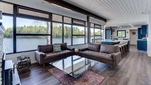 Un vaste espace de vie agrémenté d’une grande table basse en verre, de canapés en cuir et offrant une vue sur un lac grâce à de larges fenêtres.