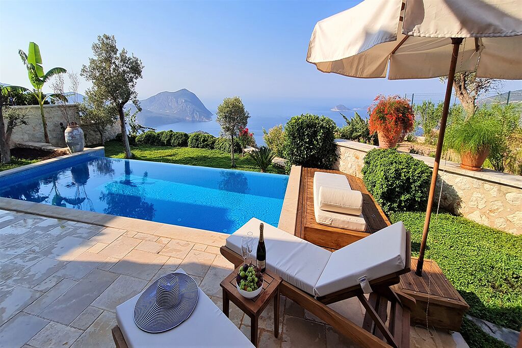 Ein Poolbereich mit Liegestühlen, einem Tisch mit Flasche und Gläsern sowie Blick auf das Meer und die Berge.