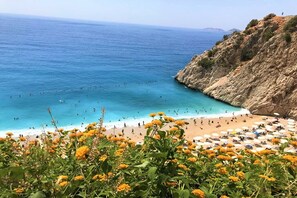 A beach with clear blue water, rocky cliffs, and a crowd of people.