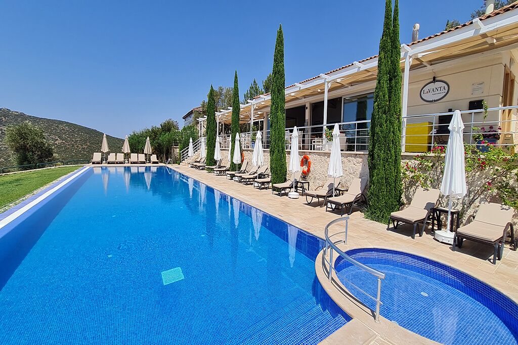 A swimming pool with lounge chairs and umbrellas, a building with a sign reading "La VANTA", and a mountainous backdrop.