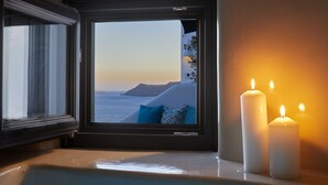 Uma banheira com vista para o pôr do sol sobre o mar, duas velas acesas e uma janela com cortinas.