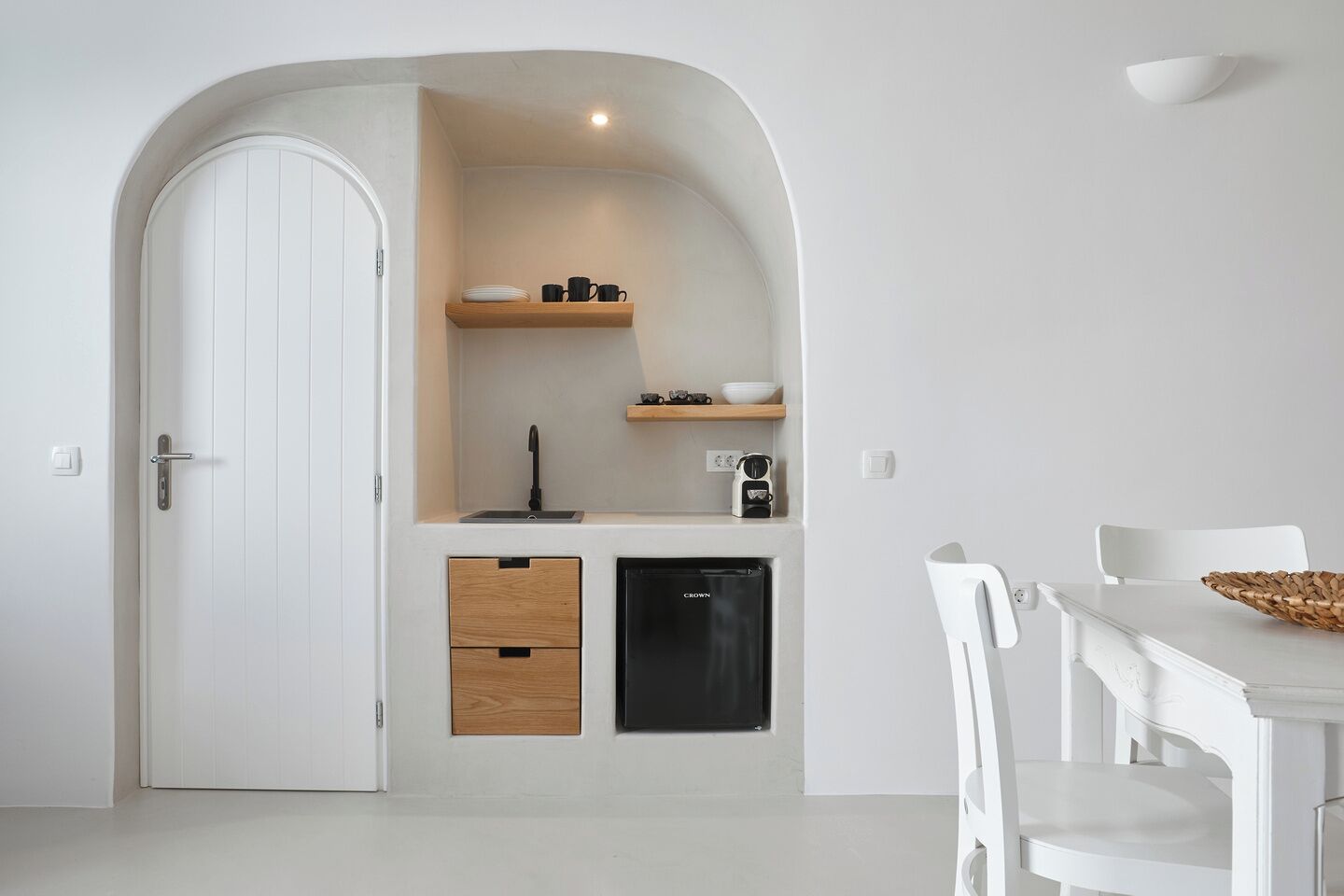A compact kitchen with a white door, wooden drawers, a black refrigerator, and a small shelf with kitchenware.