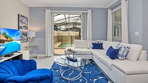 Ein modernes Wohnzimmer mit einem weißen Sofa, einem blauen Sessel und einem Glastisch. Ein Flachbildfernseher steht auf einem Schrank, und ein Glastür führt zu einem Balkon mit einem Pool.