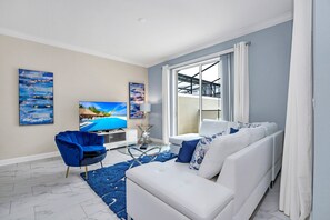 Un salon moderne avec un canapé blanc, un fauteuil bleu et une table basse en verre. Deux tableaux ornent le mur, et une grande fenêtre est agrémentée de rideaux.
