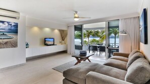 A modern living room with a large sofa, a television, and a dining area with a view of the ocean. - Frangipani Lodge 104, Frangipani Lodge 104 (Hamilton Island)