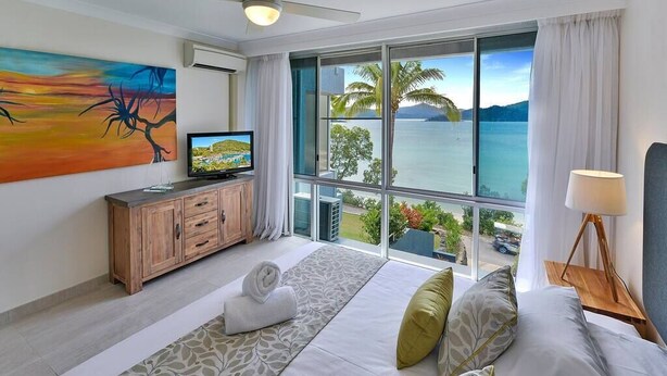 A hotel room with a large window offering a view of the sea, a bed with pillows, a TV on a wooden cabinet, and a painting on the wall. - Frangipani Lodge 104, Frangipani Lodge 104 (Hamilton Island)