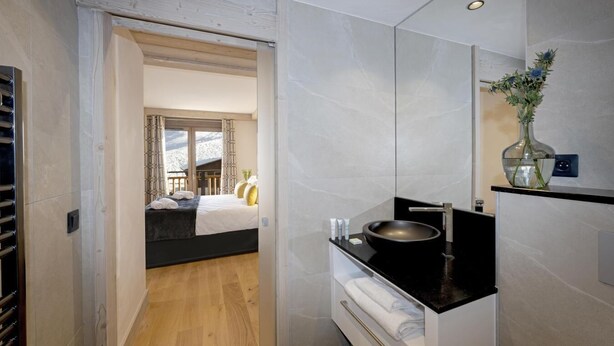 Une chambre d’hôtel moderne avec un grand lit, une salle de bain dotée d’un lavabo et d’un miroir noirs, et une vue sur la montagne par la fenêtre.