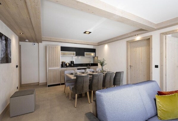 A modern kitchen with a dining area, wooden ceiling beams, and a grey sofa with a yellow pillow.