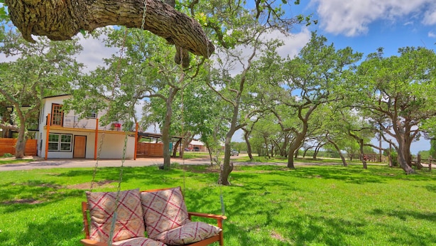 A wooden swing chair with patterned cushions in a lush green lawn, a modern building with a balcony, and a clear blue sky. - Pancho and Lefty's - Happy Days Escape. 50's Themed getaway! (Wimberley)