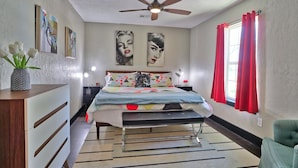 A bedroom with a bed, a ceiling fan, a dresser, a nightstand, a window with red curtains, and artwork on the walls. - Pancho and Lefty's - Happy Days Escape. 50's Themed getaway! (Wimberley)