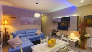 A living room with a blue sofa, a dining table with a basket of fruit, and a TV mounted on the wall. - Rosa Apartament (Palermo)