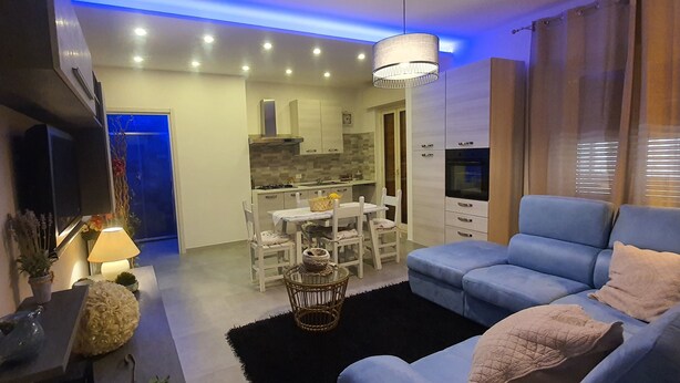 A modern living room with a blue sofa, a dining table with chairs, a kitchen in the background, and a TV mounted on the wall. - Rosa Apartament (Palermo)