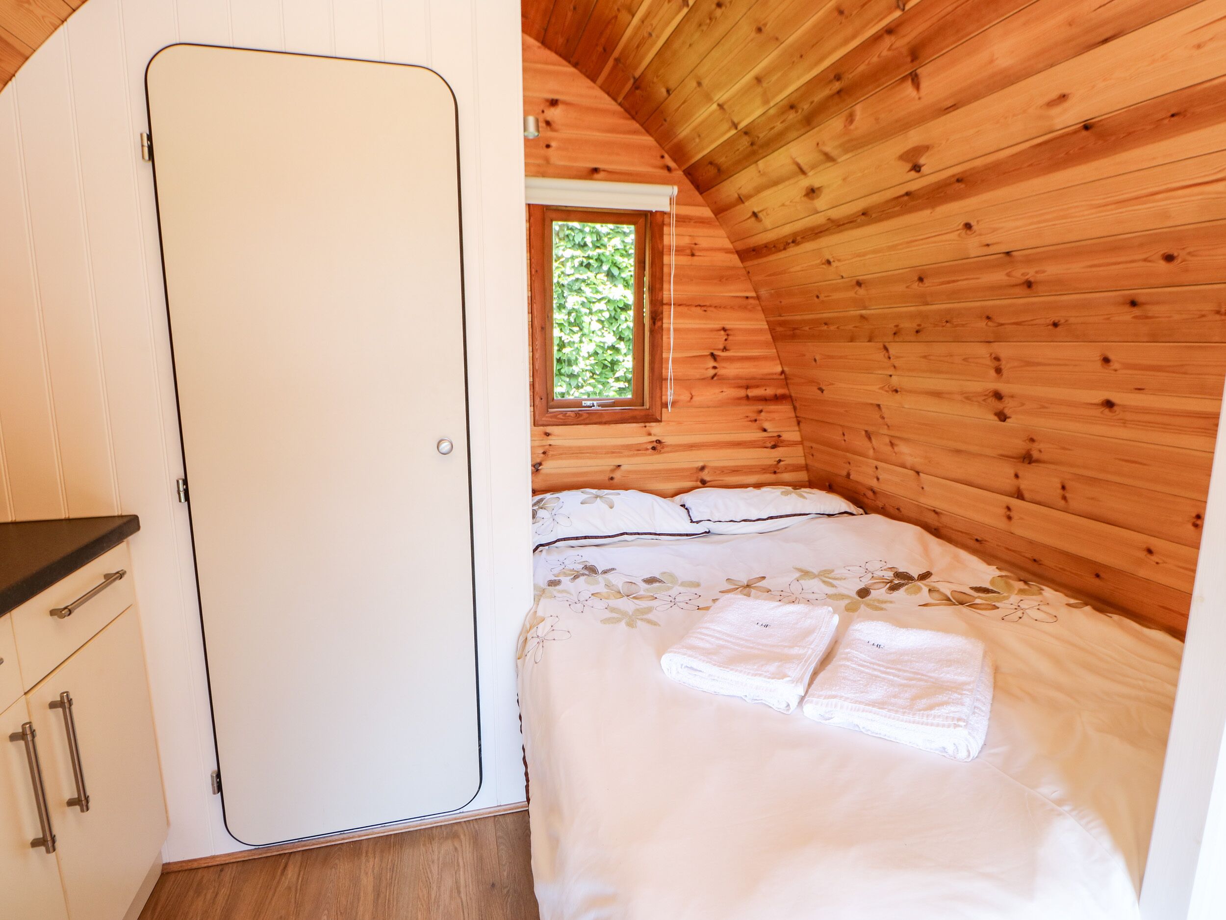 A cozy bedroom with a wooden ceiling, a bed with white linens, and a window with a view of greenery.