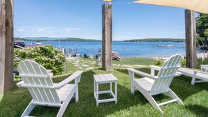 Une terrasse avec des fauteuils Adirondack et une petite table, donnant sur un lac où l’on peut voir des bateaux et un quai.