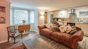 Ett mysigt vardagsrum med en brun lädersoffa, ett träbord och en röd matta. En matplats med ett träbord och stolar syns. Köket har vita skåp och mönstrade kakelplattor.