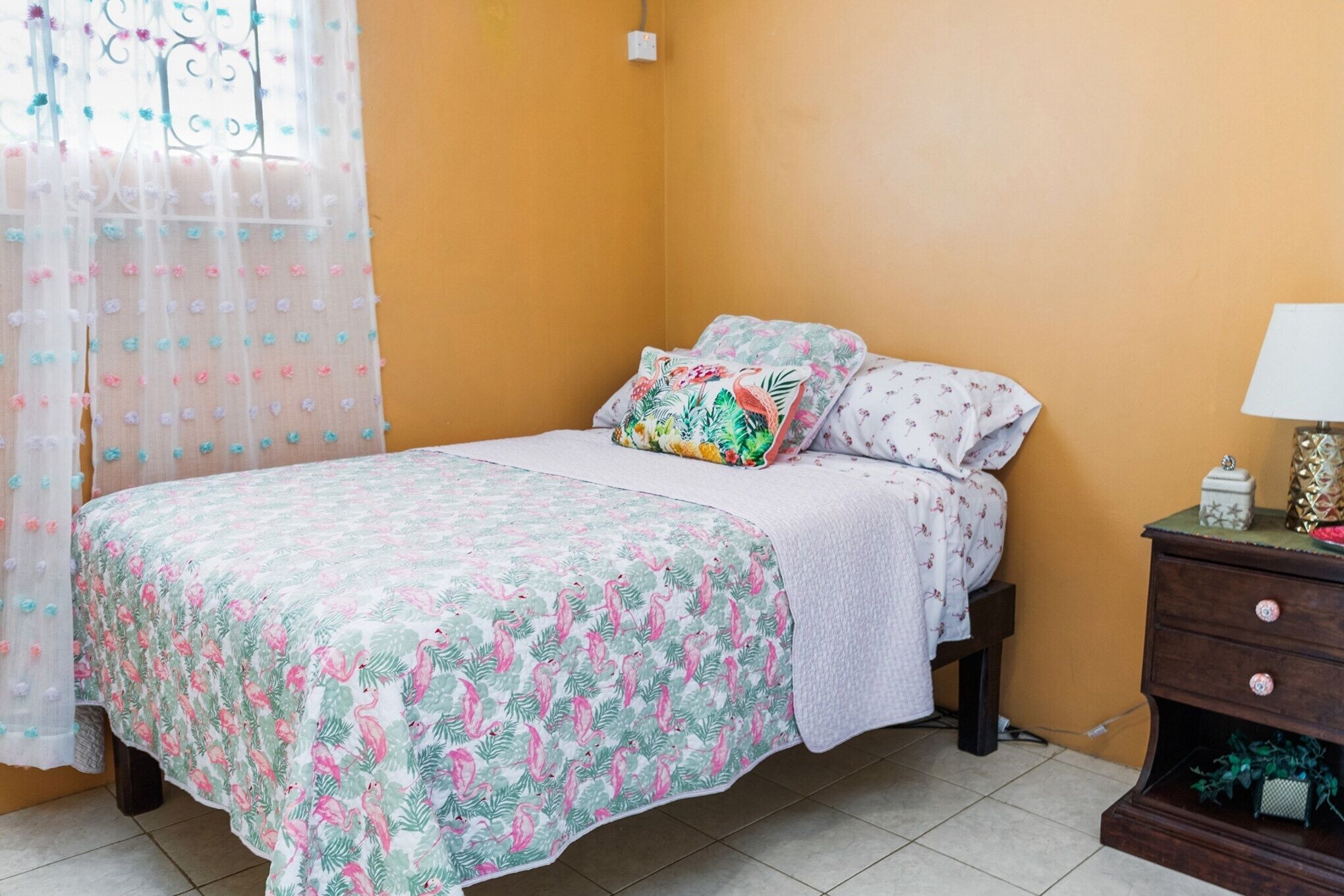Une chambre à coucher avec un lit recouvert d’une couvre-lit à motifs floraux, une table de chevet avec une lampe et une fenêtre avec un rideau.
