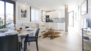Cozinha moderna com armários brancos, área de jantar com mesa redonda e sala de estar com sofá.