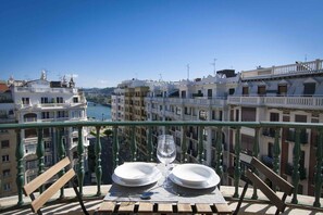 A balcony table with two plates and a wine glass, overlooking a cityscape with buildings and a river.