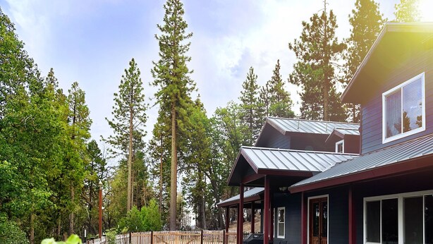 A house with a covered patio, a gravel driveway, and tall trees in the background.