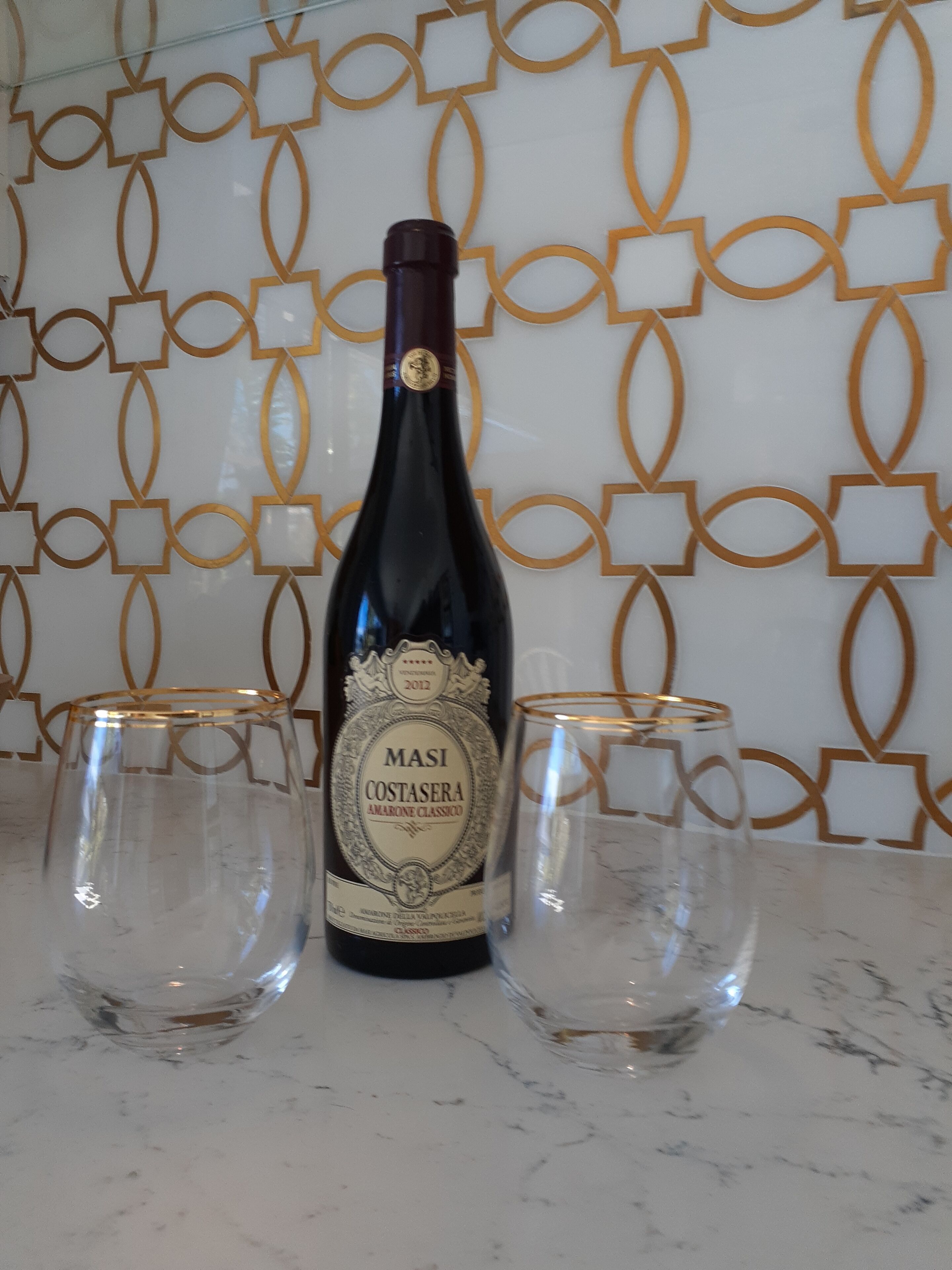 A bottle of Masi Costasera Amarone Classico wine with two empty glasses on a marble surface.