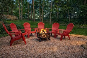 A fire pit with four red chairs arranged around it, set on a gravel surface with a grassy area and trees in the background.