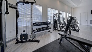 Un gimnasio con varios equipos de ejercicio, ventanas amplias y un banco.