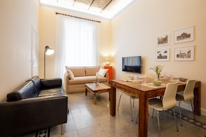 Una sala de estar con un sofá, una mesa de centro, una mesa de comedor con sillas, un televisor y cuadros en la pared.