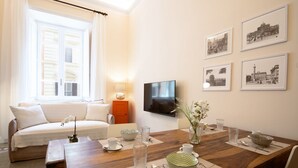 Un salon comprenant un canapé, une table à manger dressée avec de la vaisselle, un téléviseur fixé au mur et des tableaux encadrés.