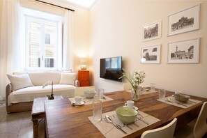 A living room with a sofa, a dining table set with glassware, a TV mounted on the wall, and framed pictures.