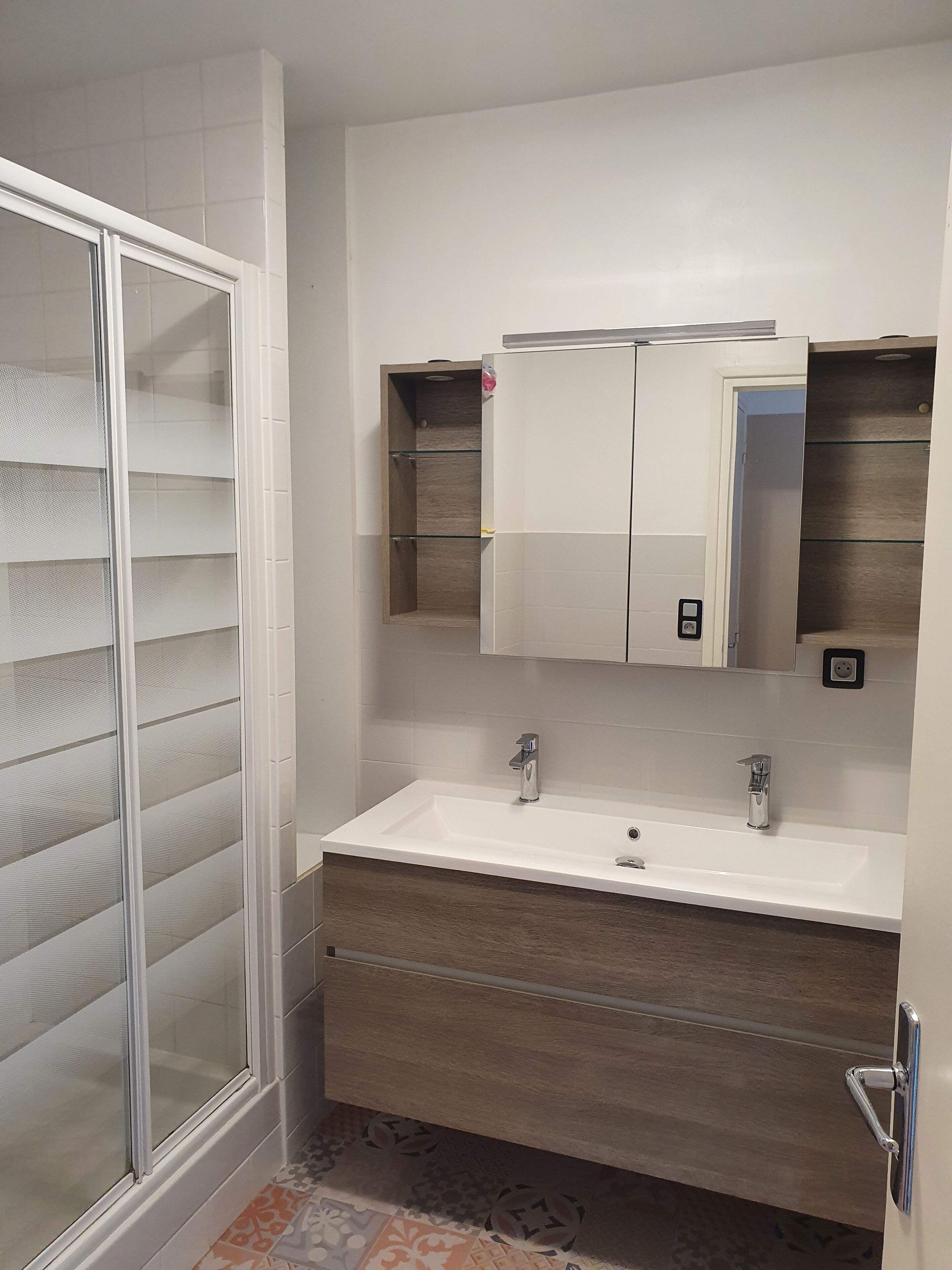 A bathroom with a glass shower enclosure, a white sink with two faucets, and a wooden vanity.