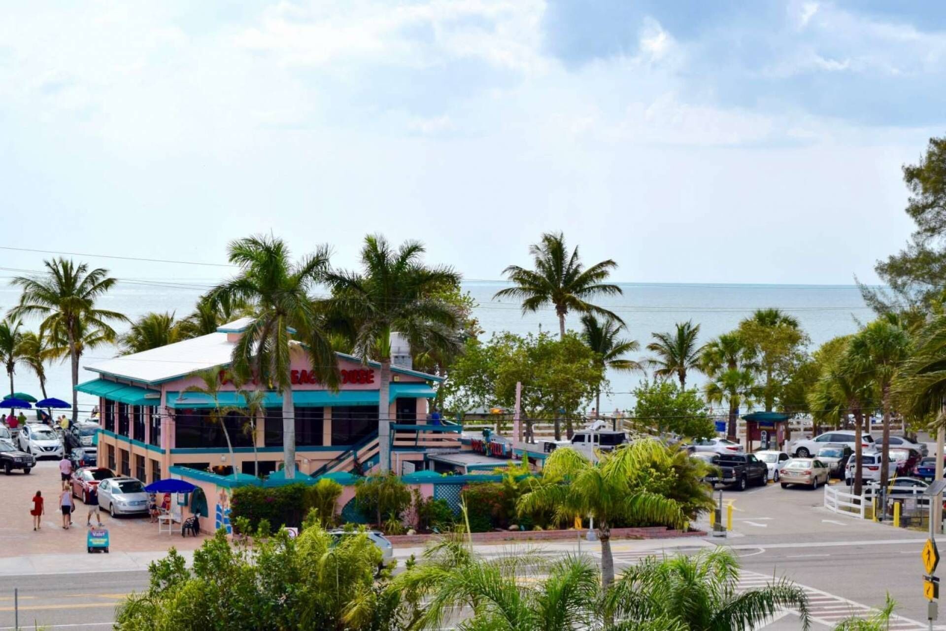 A hotel with a white facade and blue accents, located near a beach with palm trees and parked cars.