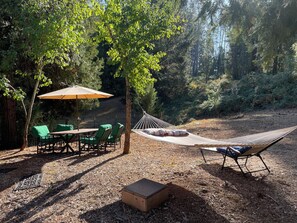 A hammock, green chairs, a table, and an umbrella in a forested area.