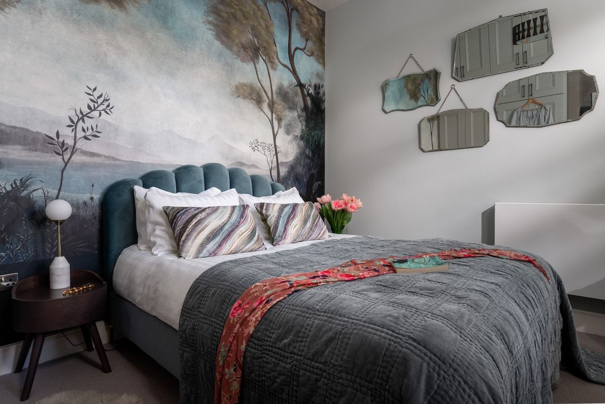 Een slaapkamer met een bed, nachtkastje, lamp, schilderij en wandopbergruimte.