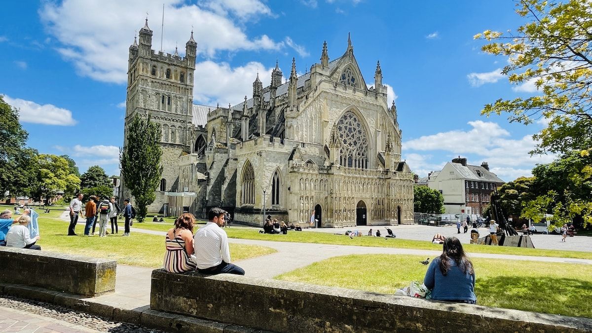 En historisk katedral med ett stort rosettfönster och två tvillingtorn, omgiven av en grön innergård där människor sitter och promenerar.