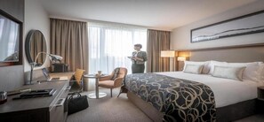 Een hotelkamer met een bed, een bureau met een laptop, een stoel, een klein tafeltje met een kopje en een staande lamp.