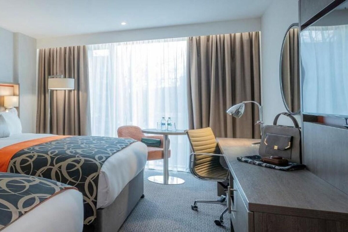 Ein Hotelzimmer mit zwei Betten, einem Schreibtisch, einem Sessel und einem großen Fenster mit Vorhängen.