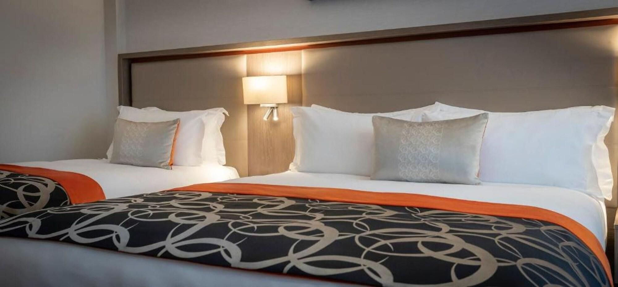 Habitació d'hotel amb dos llits, cadascun amb roba de llit blanca i coixins decoratius. Hi ha una làmpada penjada a la paret entre els llits.