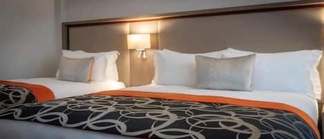 A hotel room with two beds, each with white linens and decorative pillows. There is a wall-mounted lamp between the beds.