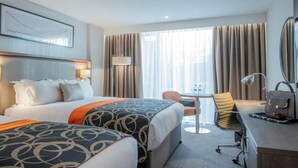 Une chambre d’hôtel avec deux lits, un bureau, une chaise, une lampe et une grande fenêtre avec des rideaux.