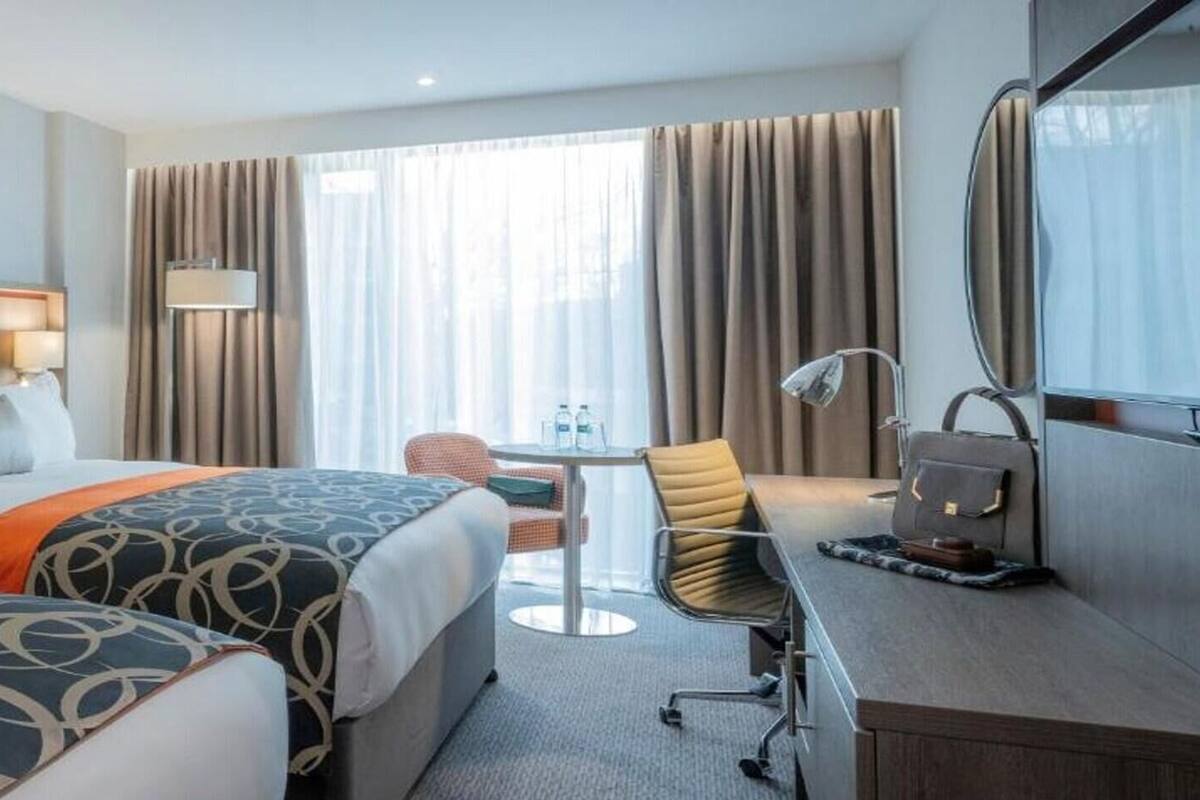 Habitación de hotel con dos camas, un escritorio, una silla, una lámpara y un ventanal grande con cortinas.