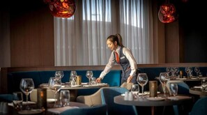 Ein Restaurant mit gedeckten Tischen, einem Kellner, der sich um eine Gruppe kümmert, und einem modernen Ambiente.